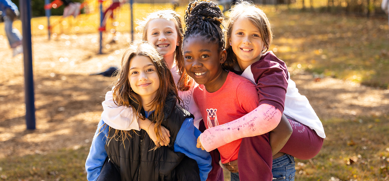 Four young girls smile happily together outdoors on a playground, two of them giving piggyback rides to the other two.