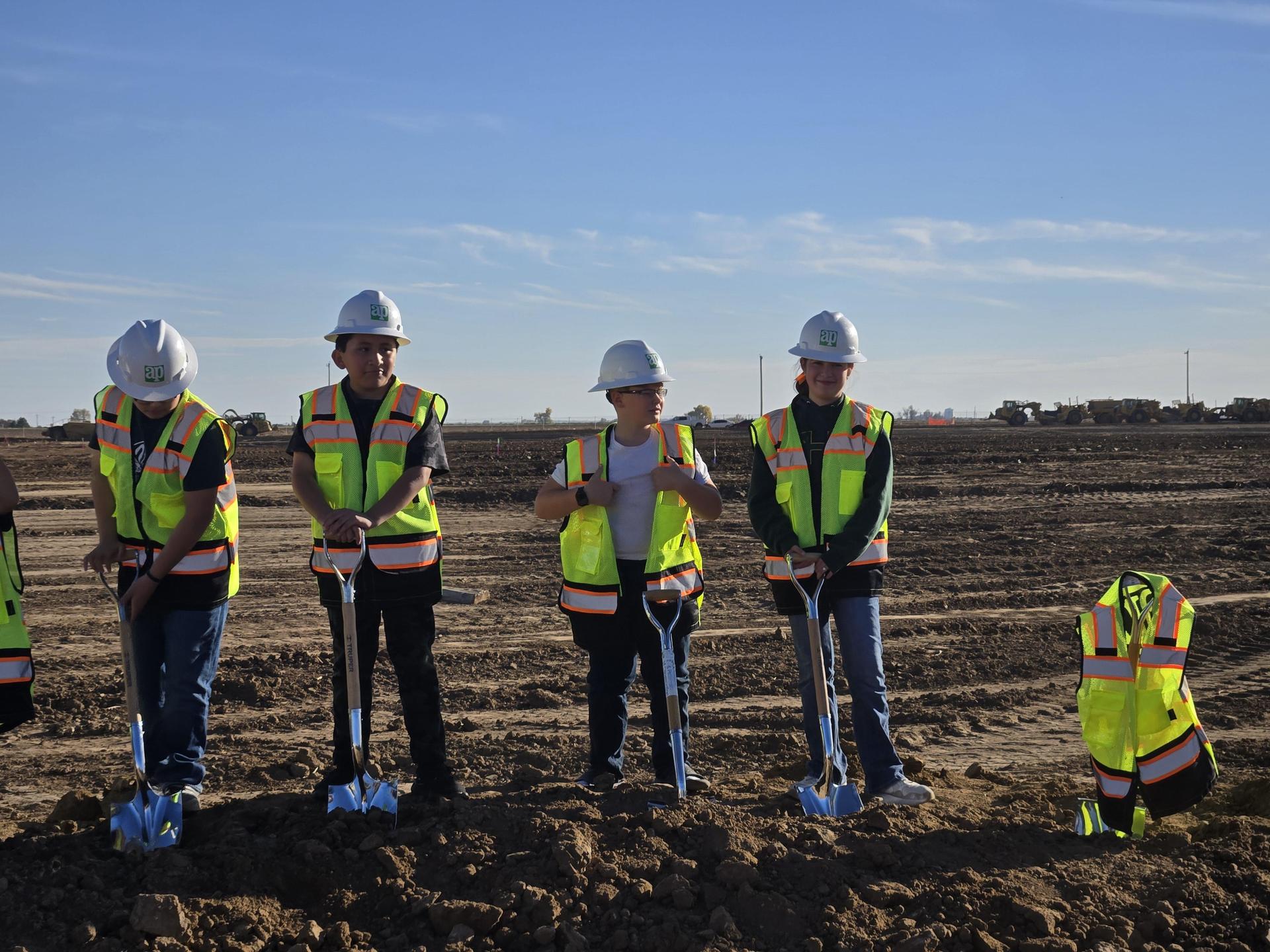 Children in hard hats and vests stand with shovels on prepared soil at a construction site.