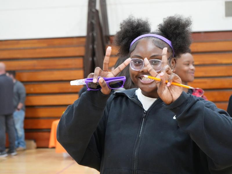 Girl making a peace sign with glasses, holding a pencil in a gym setting.