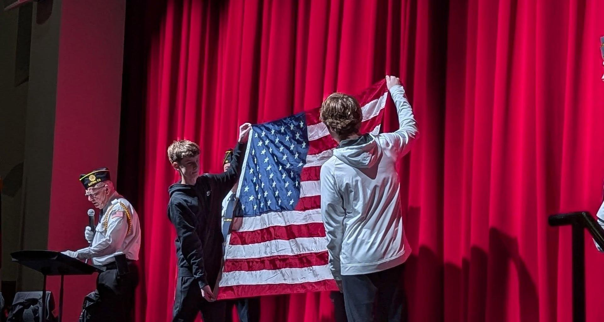 Two boys hold an American flag in front of a man at a podium with red curtains.