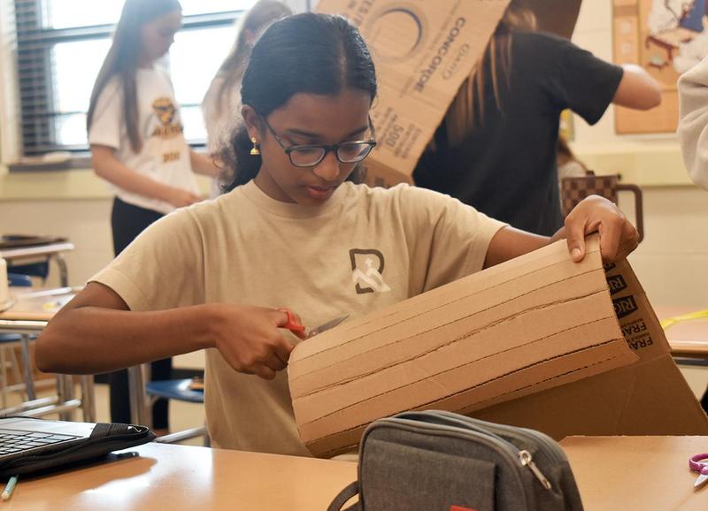 Girl with glasses cuts cardboard in a classroom setting.