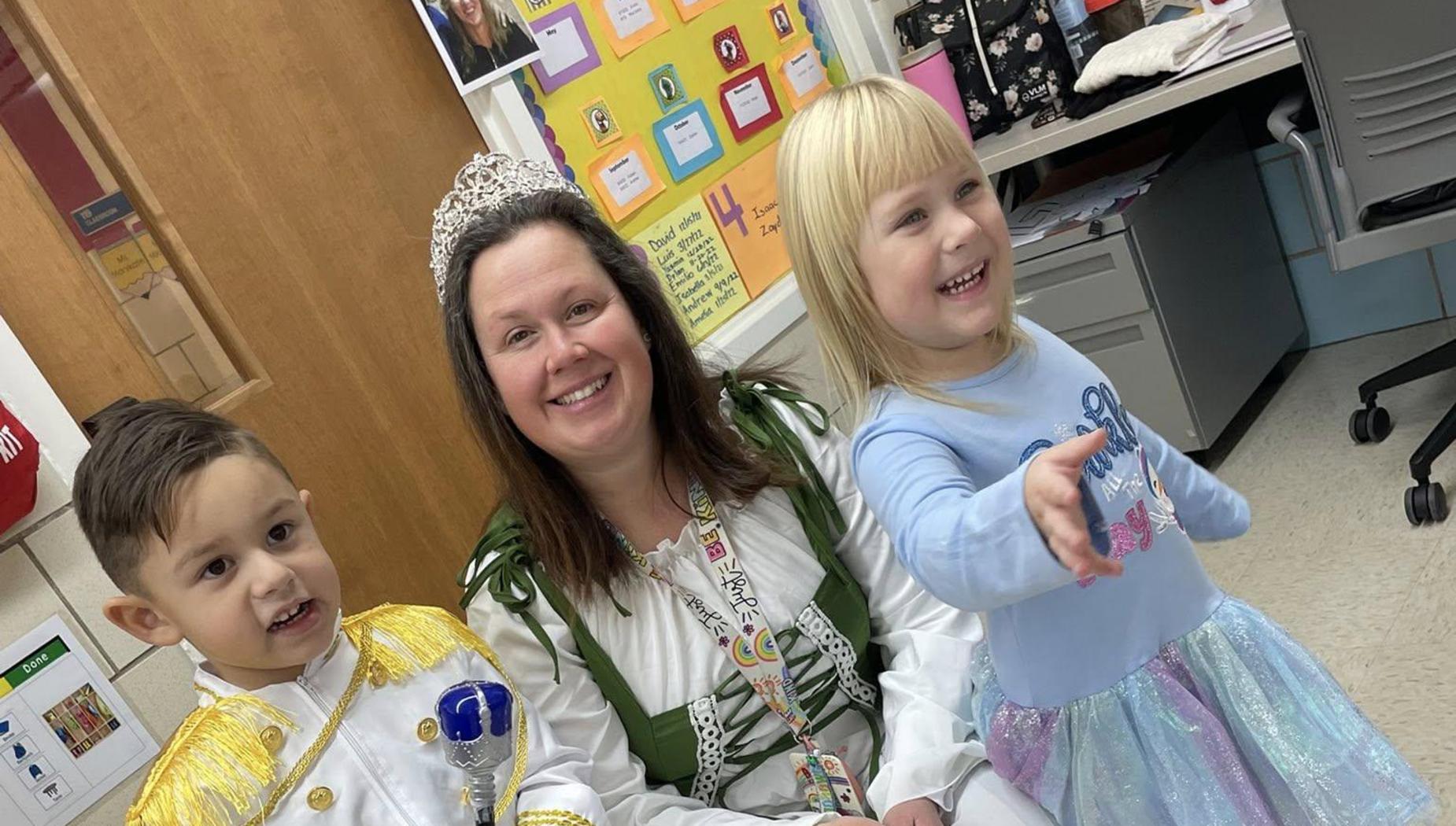 A teacher and two children in costumes, smiling and sitting together in a classroom.