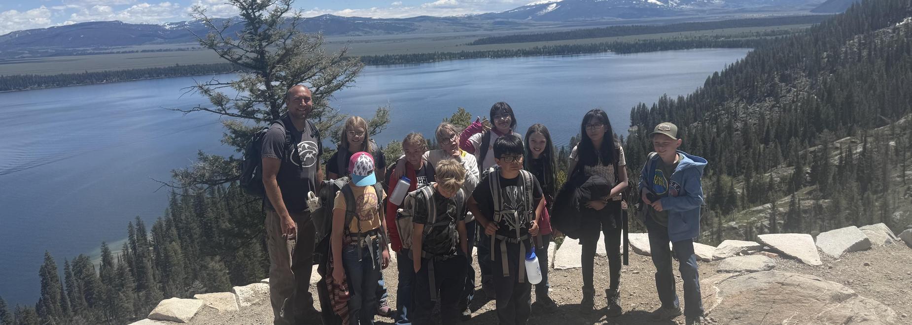 Group of children and adults posing with a scenic view of a lake and mountains.