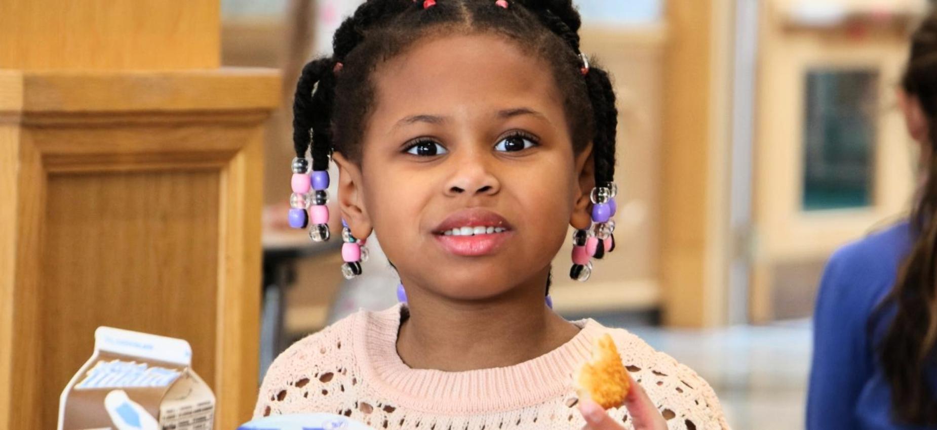 Young girl with braided hair holds a snack in a school cafeteria.