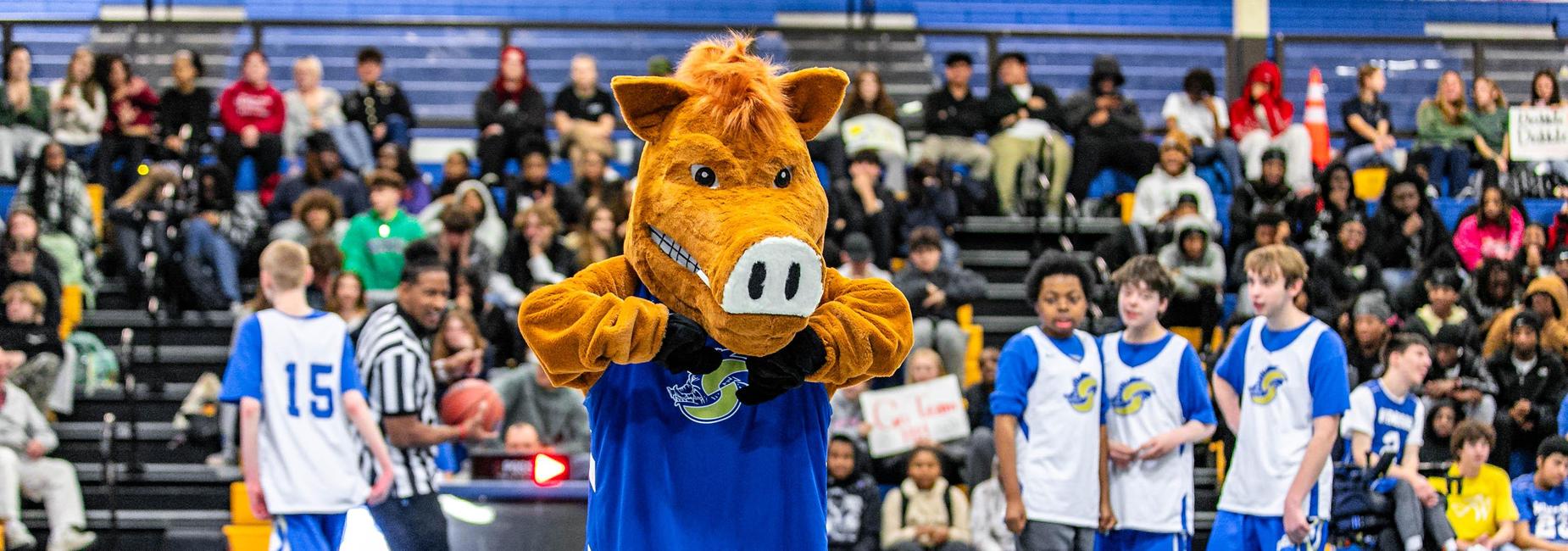 A crowd watches a basketball game as a mascot in a brown costume stands on the court.
