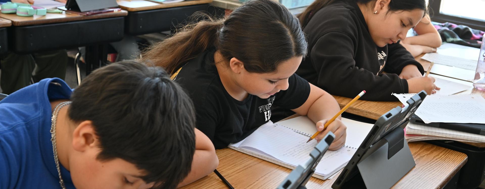 Students concentrate on their work with tablets and notebooks at a classroom table.