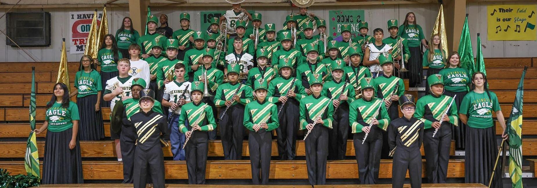 A marching band group in green uniforms, holding instruments and flags, arranged in rows.