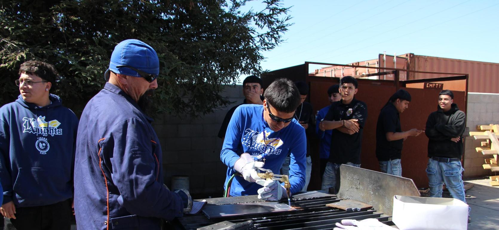 A group of students engaged in welding practice outdoors, with one focused on a task.