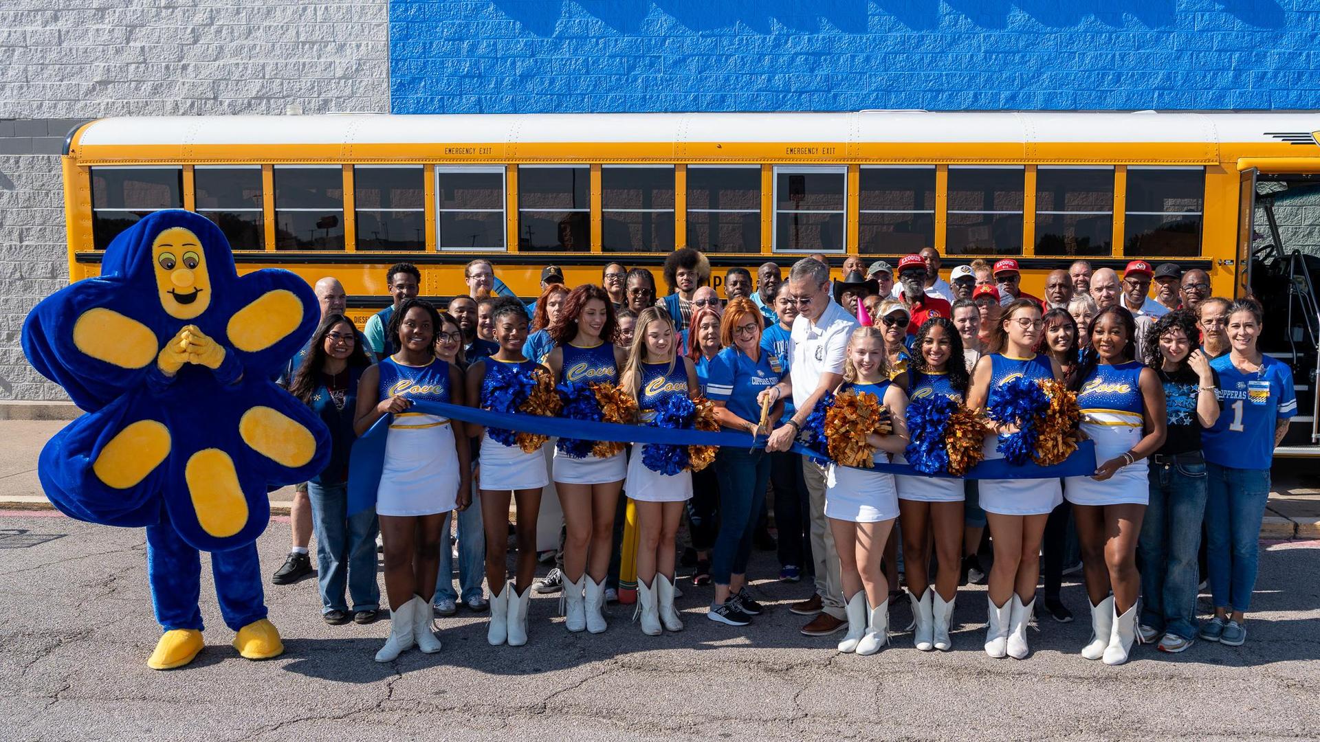 A large group of people with a mascot in front of a school bus, celebrating.