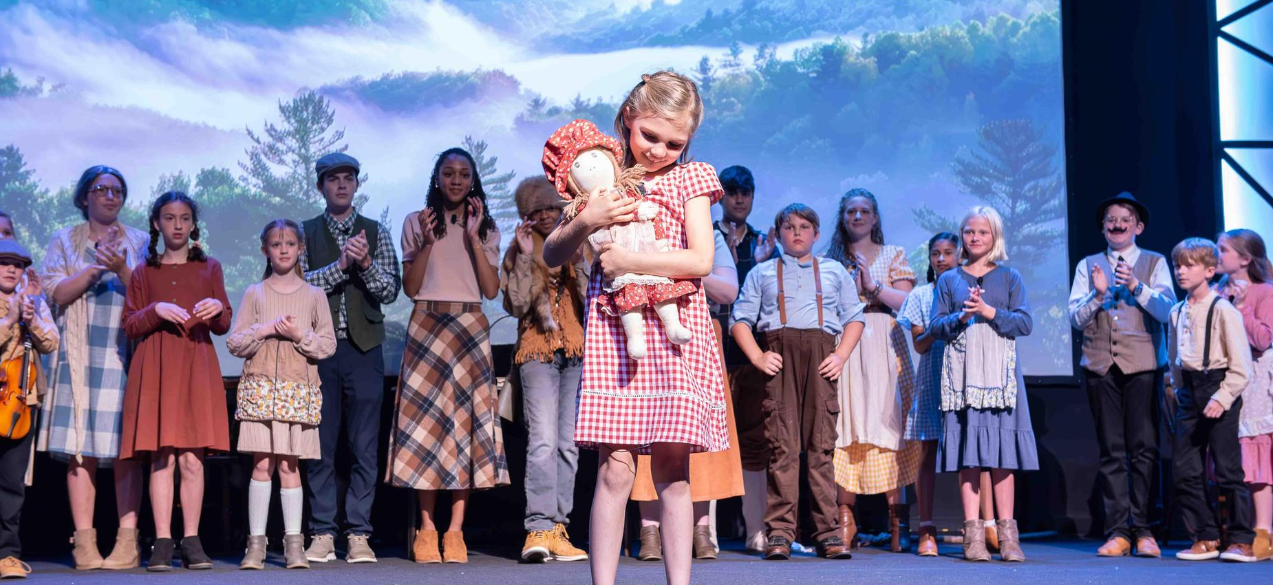 Child in a red checkered dress holds a doll on a stage with a large group applauding.