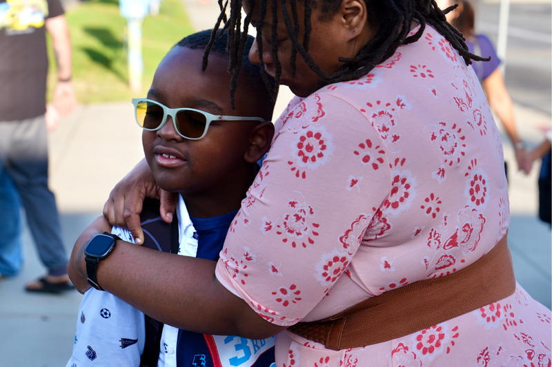 A Black woman embraces her eight-year-old Black son at the entrance of a school.