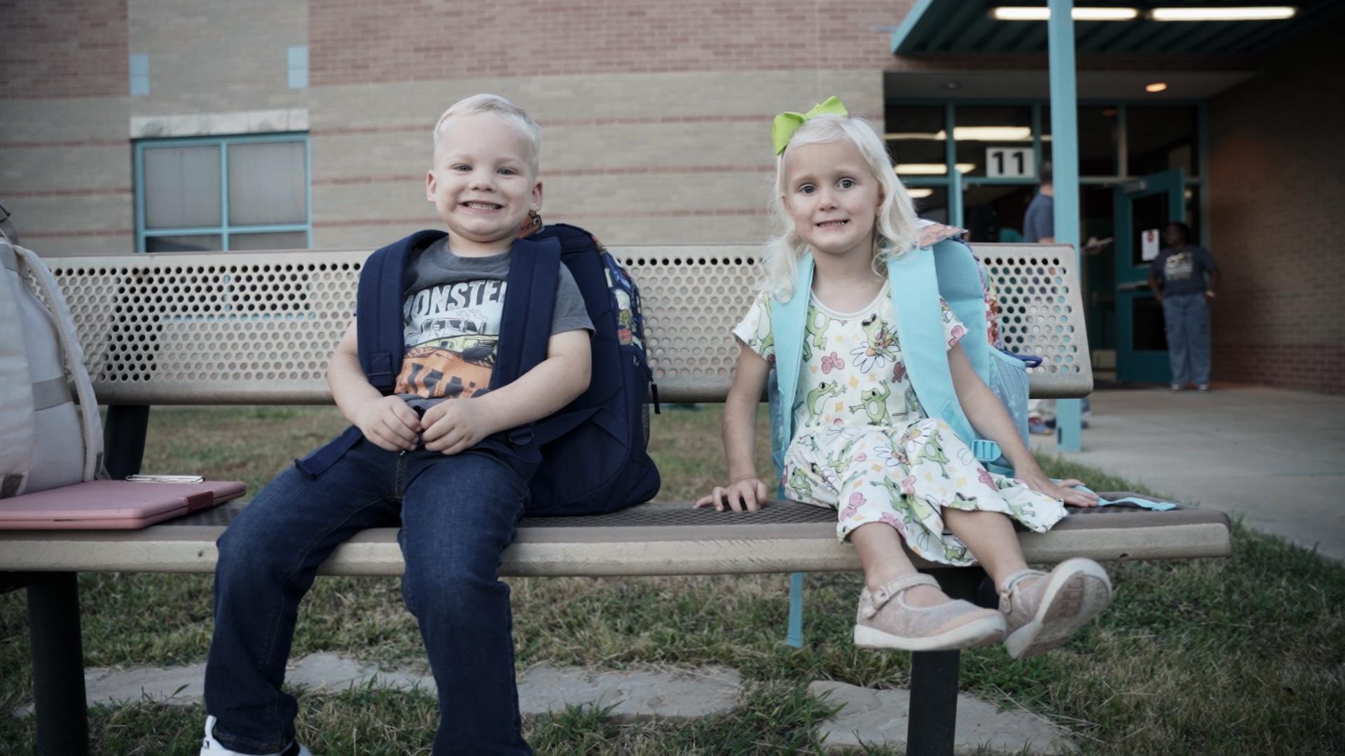 Two children sitting on a bench, smiling and wearing backpacks.
