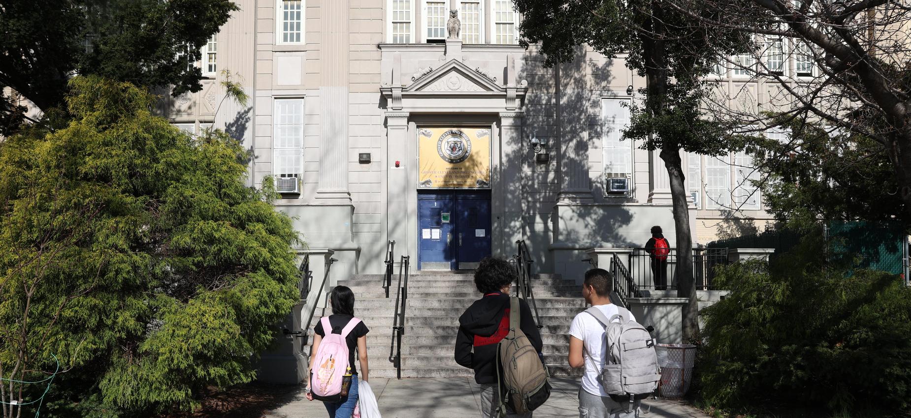 Three students approaching a school entrance with greenery around.