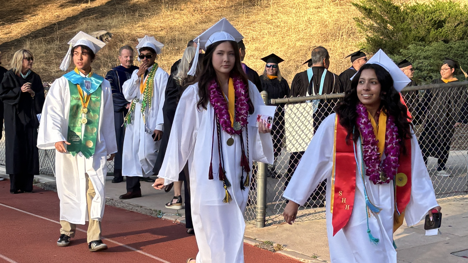 Three female graduates in white gowns with leis walking on a track.