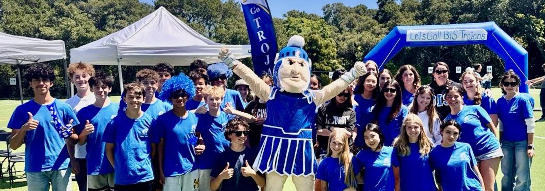 Group of students in blue shirts with a mascot at an outdoor event.