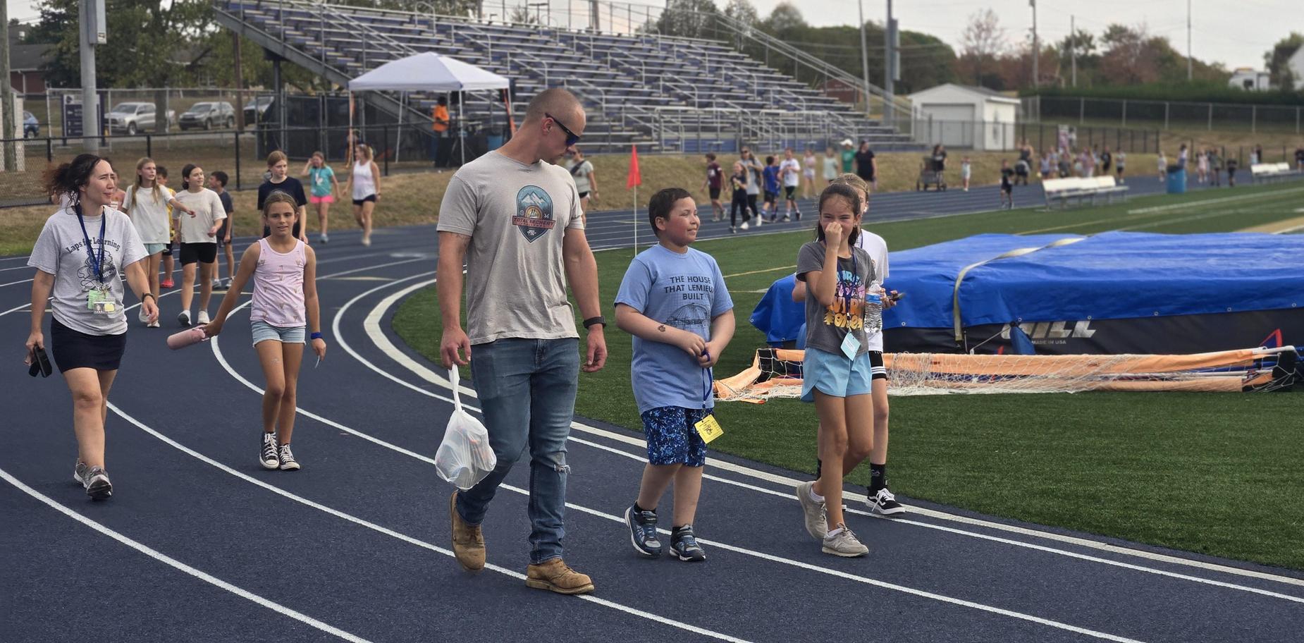 Group of people walking on a track, some chatting, with bleachers in the background.