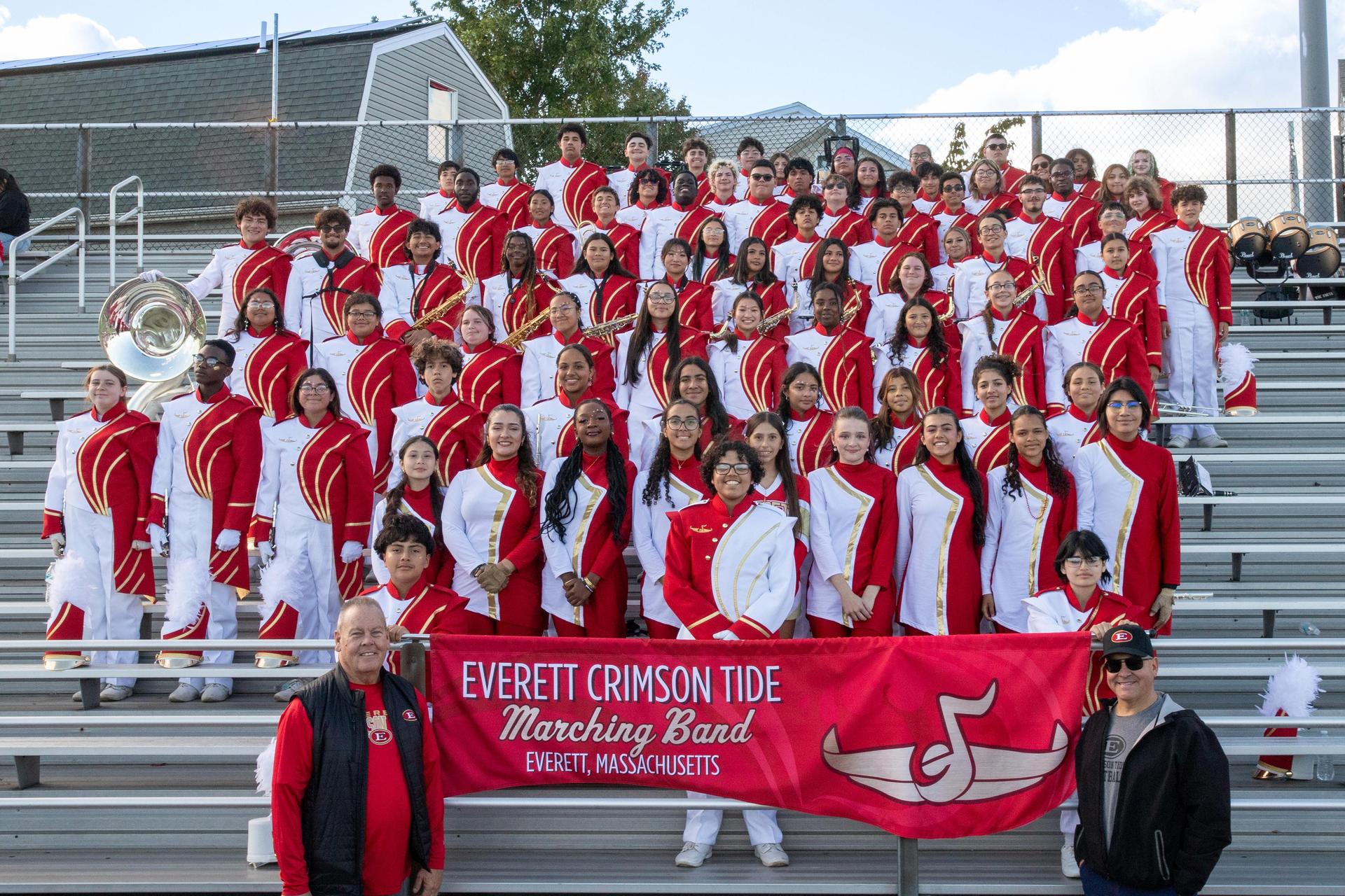 Large ensemble of musicians in uniforms posing on bleachers with a banner.