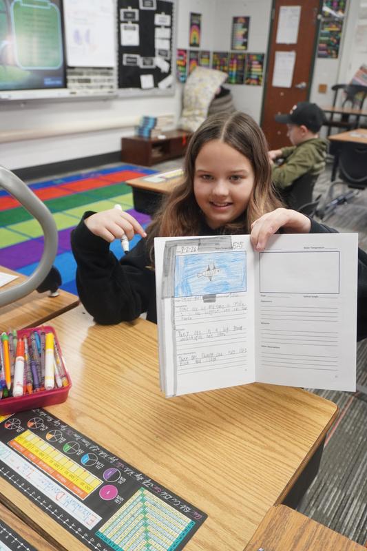 A student shows a picture she drew and colored in her booklet.