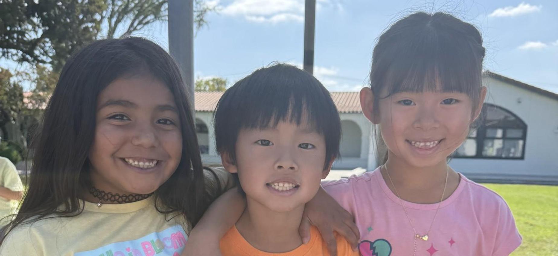 Three smiling children posing together outdoors in colorful t-shirts.
