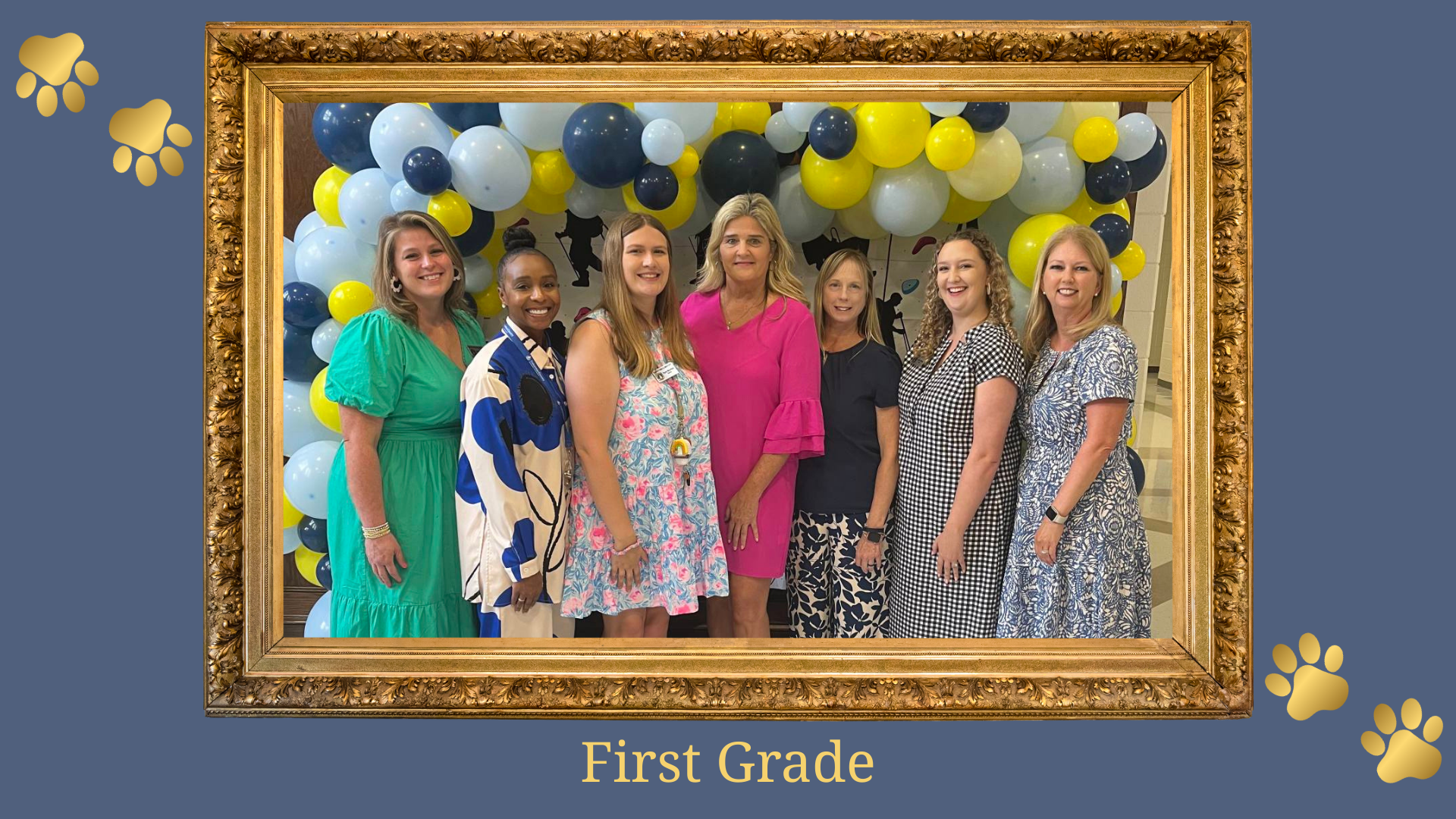 Seven women pose together in front of a colorful balloon arch labeled 'First Grade'.