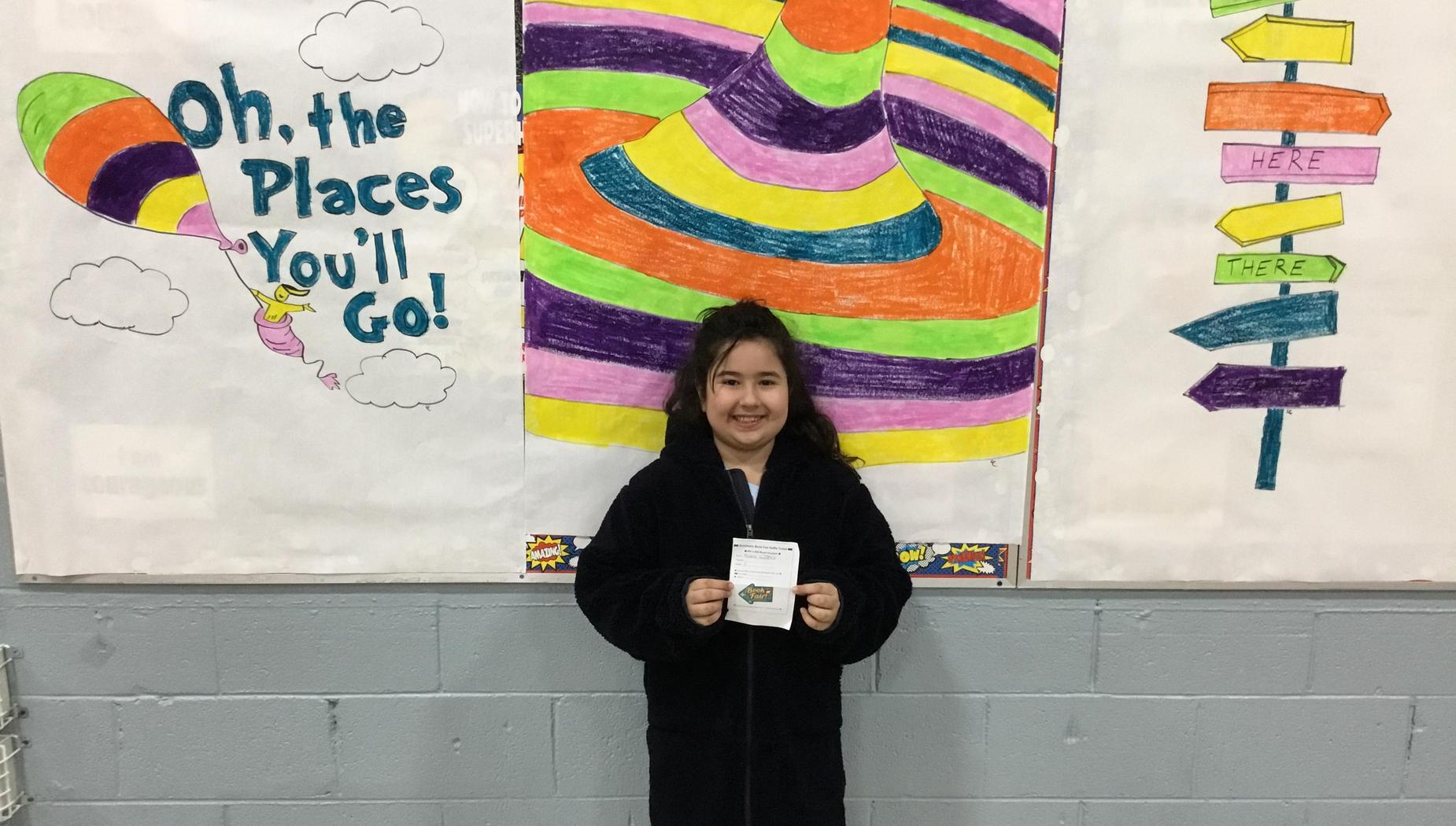 Child in a black robe holding a certificate in front of a colorful mural and signs.