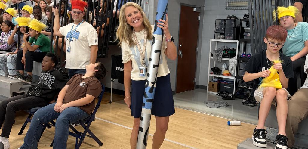 A smiling woman holds a large foam pole, surrounded by students in a school gym.