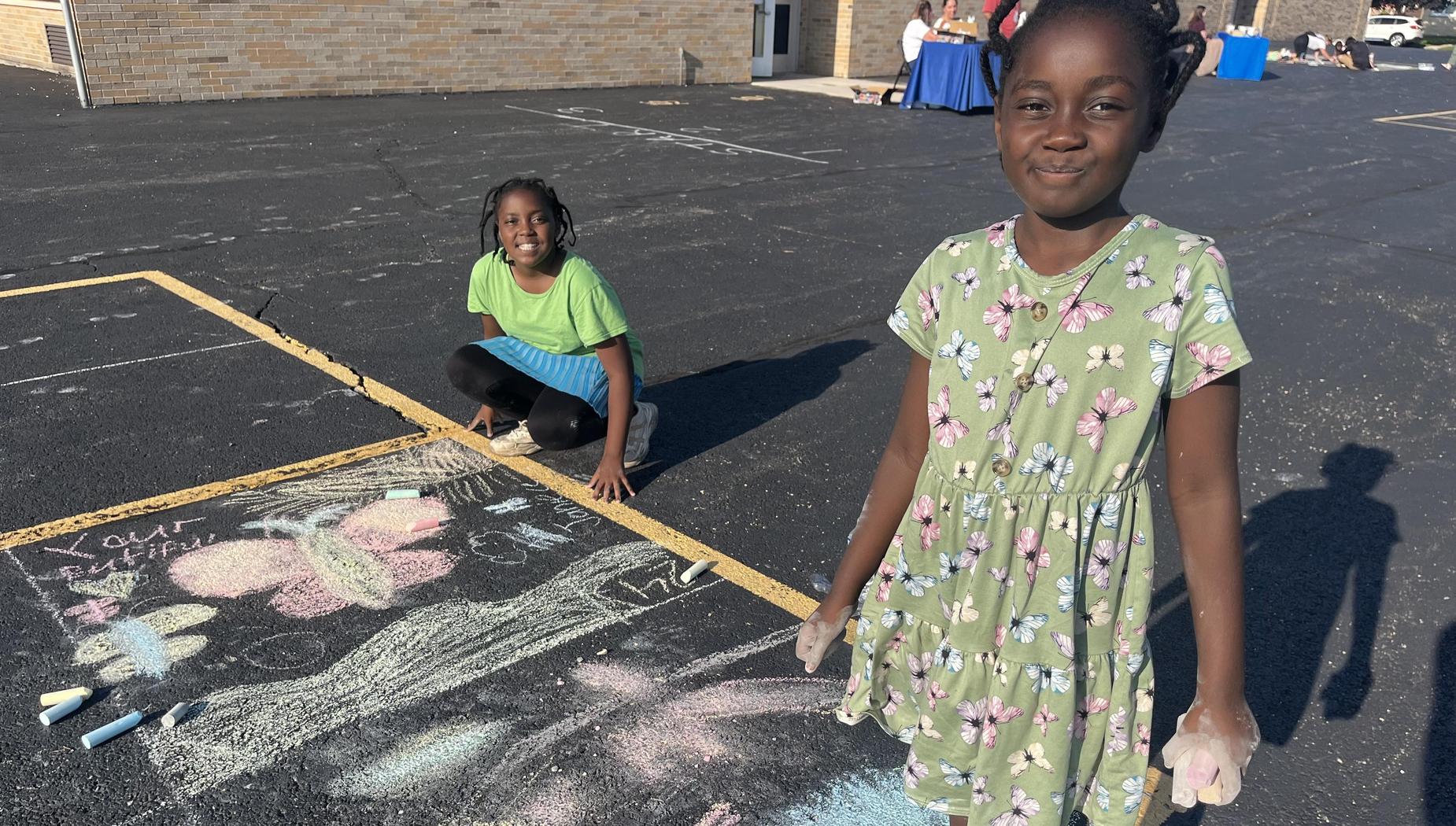 Two girls drawing with chalk on a playground, smiling at the camera.
