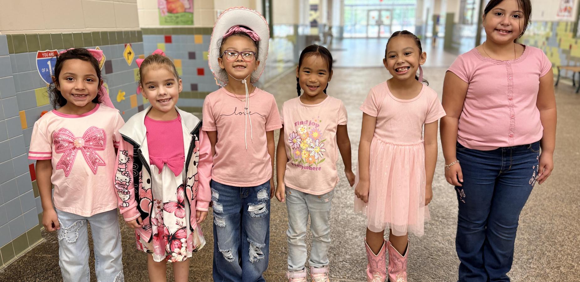 Six girls dressed in pink stand together, smiling in a school hallway.