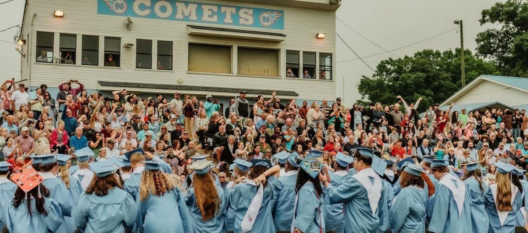 Graduates in blue gowns facing a crowd celebrating at a graduation ceremony.