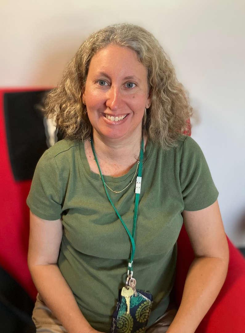 A smiling woman with curly hair, wearing a green shirt, seated in a red chair.