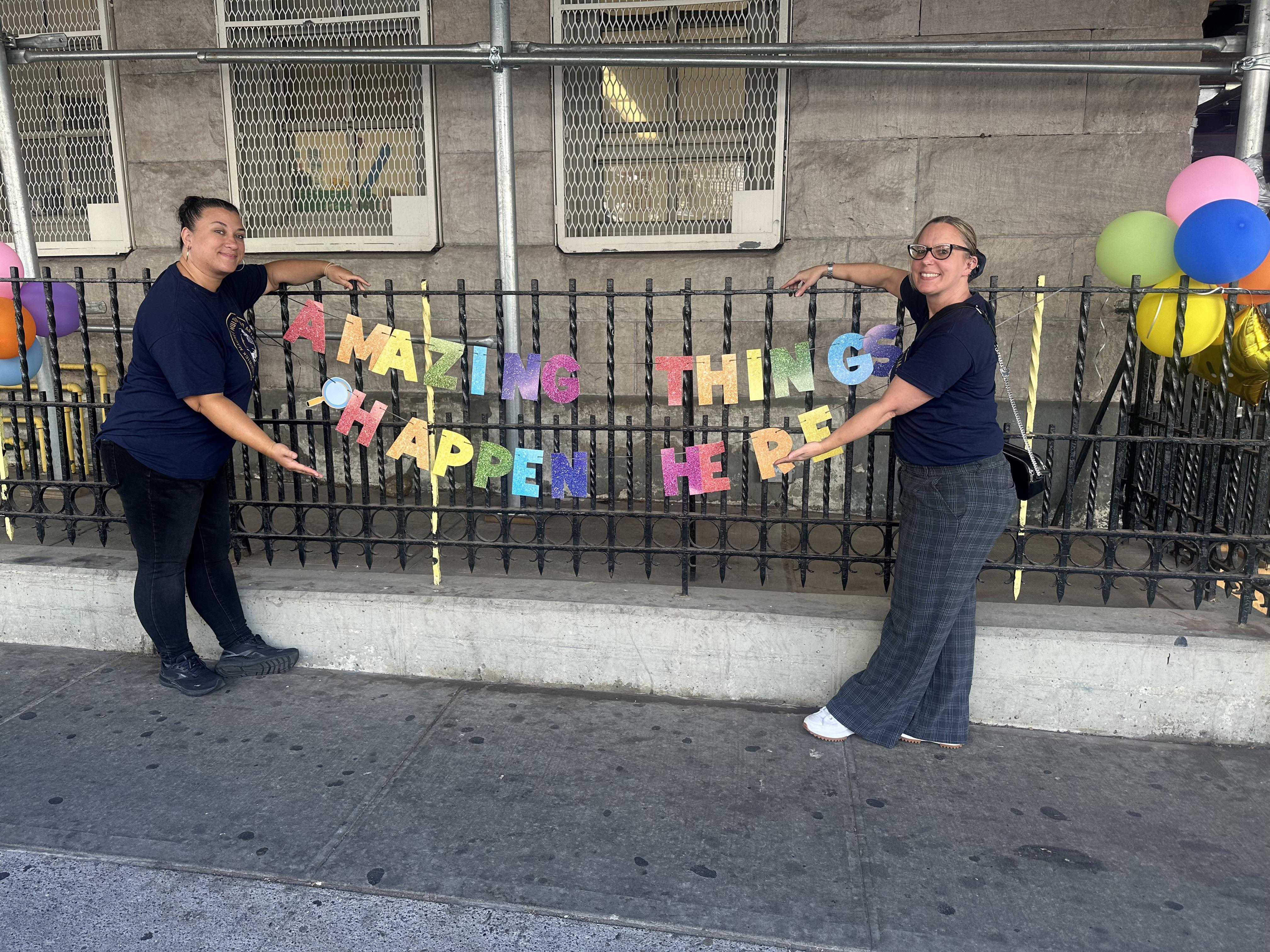 two teachers pointing to a school banner