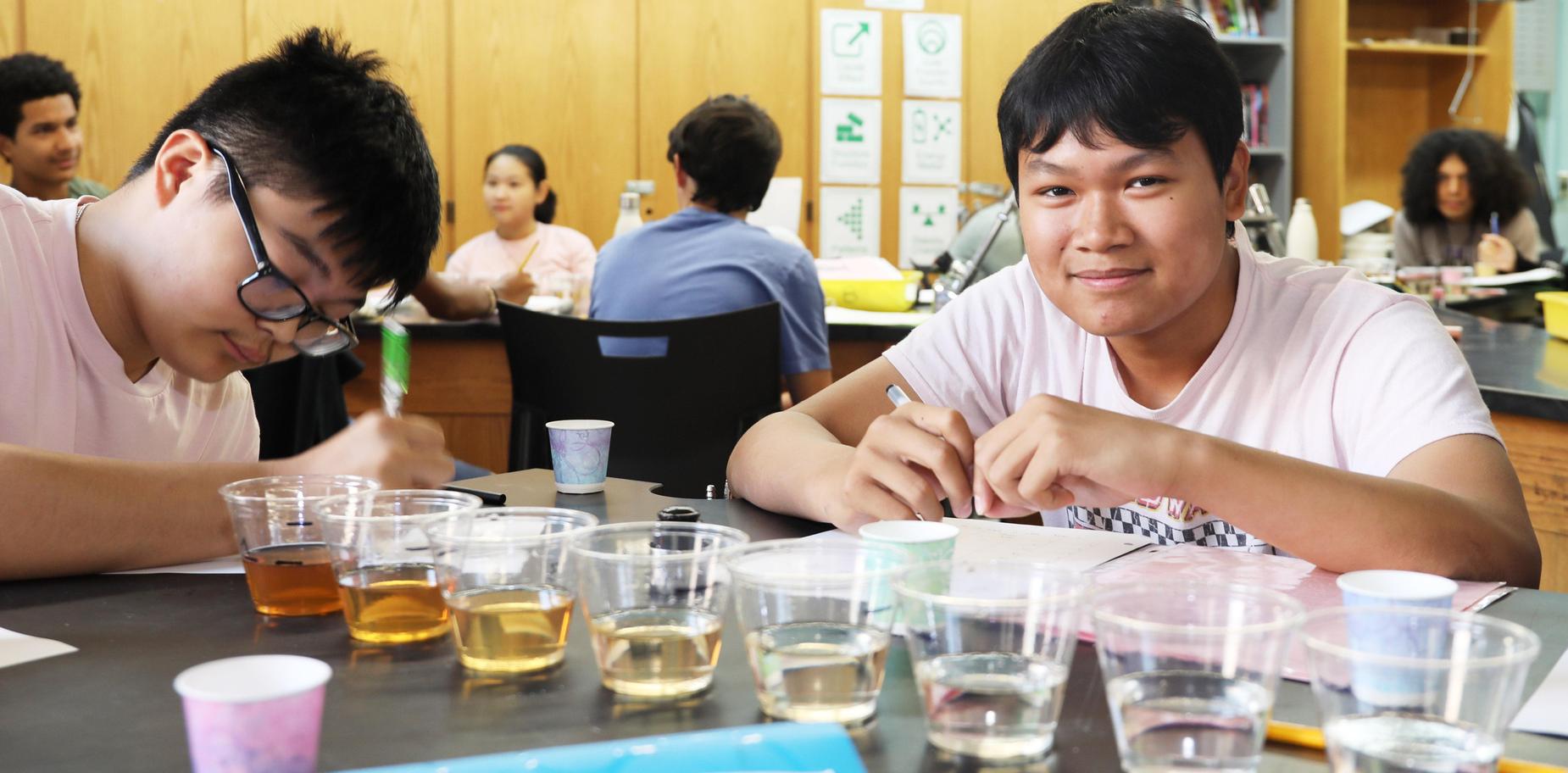 Two students working on a project at a laboratory table with multiple cups of liquids.
