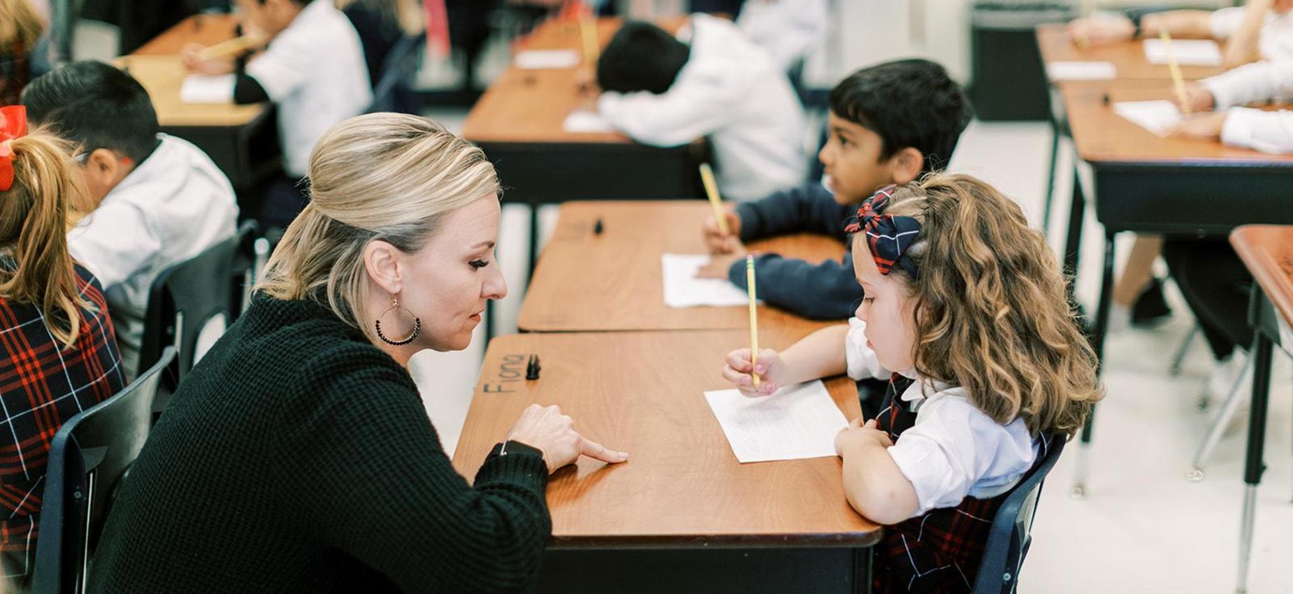 Teacher kneels beside a young student to help with her writing