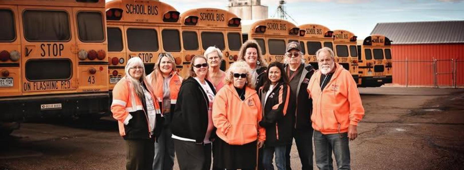 Group of school staff posing in front of parked yellow school buses.