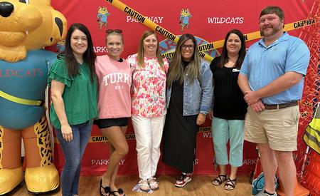 Six individuals smile together in front of a red backdrop with a large cat character and caution tape.