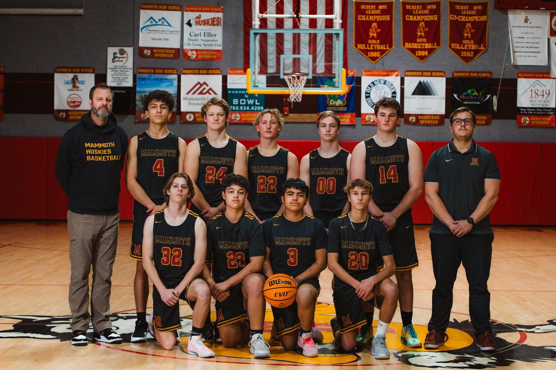 Basketball team in jerseys, posed on court with a hoop and banners in the background.