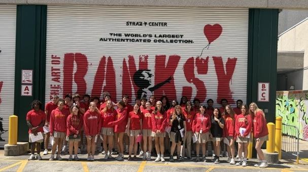 25 students in red CCC uniforms smile in front of Banksy mural