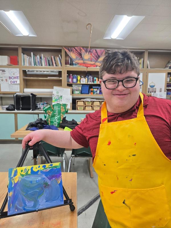 A student proudly holding up a colorful canvas on an easel.