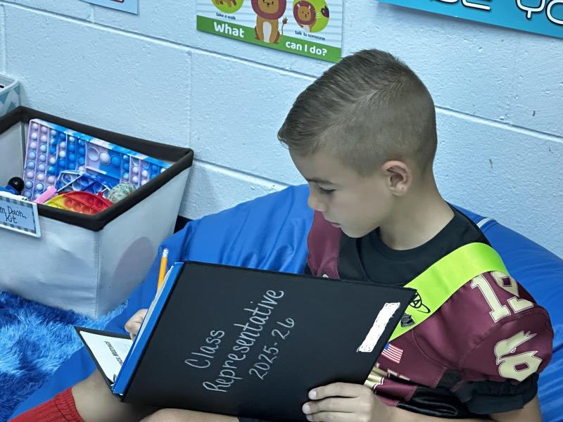 Boy sitting on a bean bag writing in a notebook labeled 'Class Representative 2025-26'.