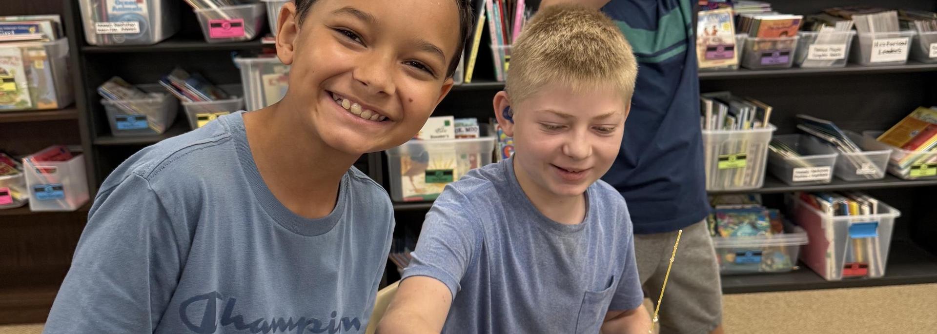 Two boys smiling while working on a project in a classroom full of books.