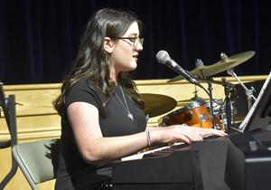 Girl in glasses playing piano and singing with drums in the background.