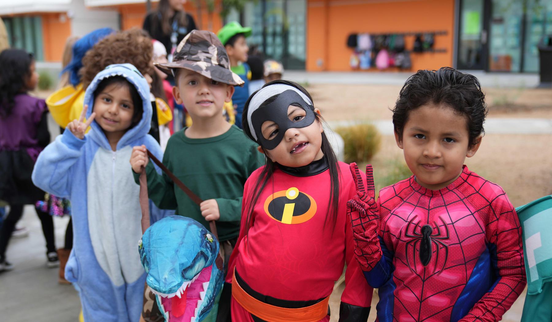 Children in costumes, including superheroes and a dinosaur, smiling in line.