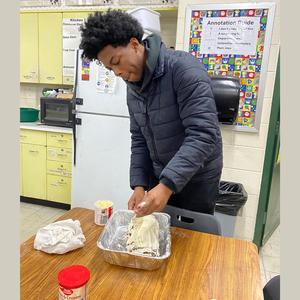 A student prepares a cake in the kitchen, adding frosting to it.