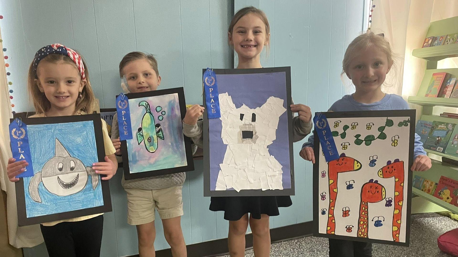 Four children proudly display their art projects, each holding a framed piece.