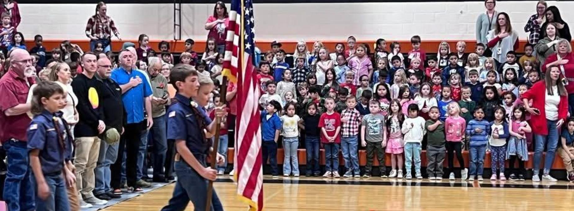 A group of children and adults standing for the national anthem in a gymnasium.