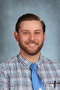 Smiling male teacher in a checked shirt and blue tie against a gray background.