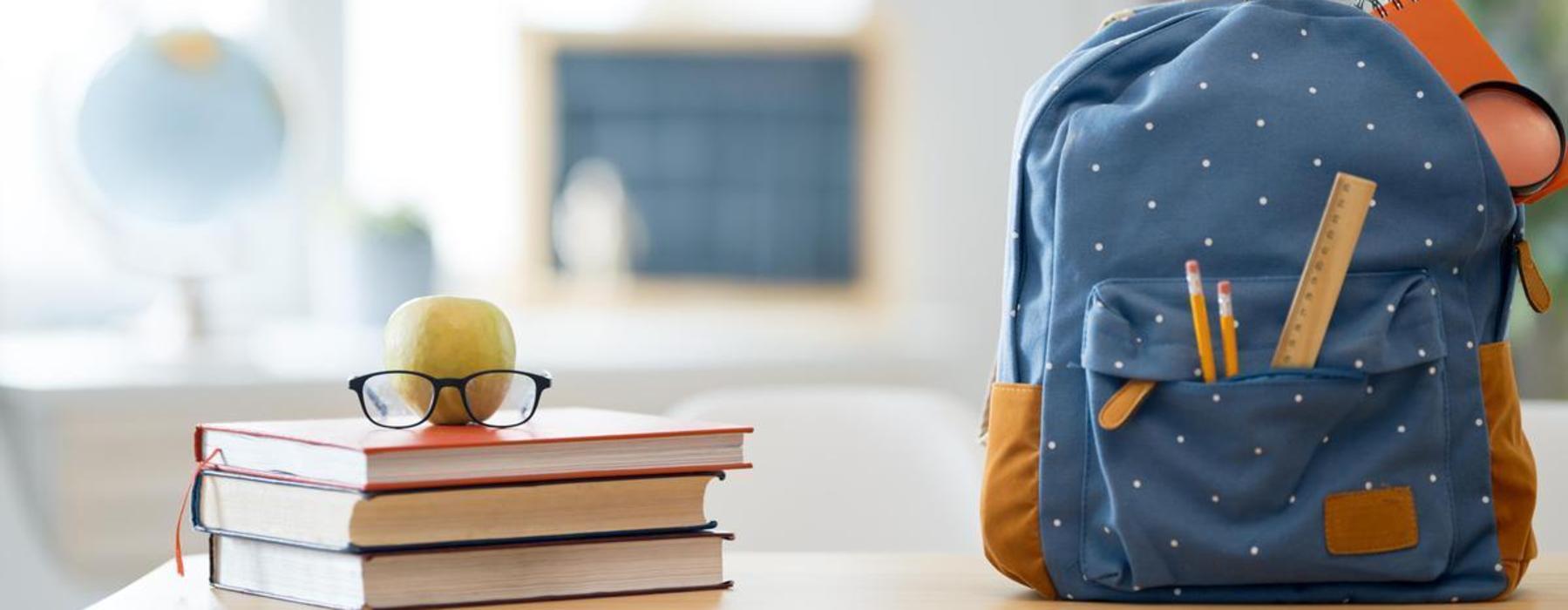 Blue backpack beside an apple, glasses, and books in a study area.