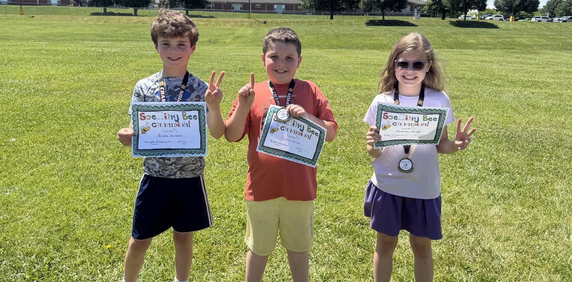 Three children displaying spelling bee certificates outdoors on a sunny day.