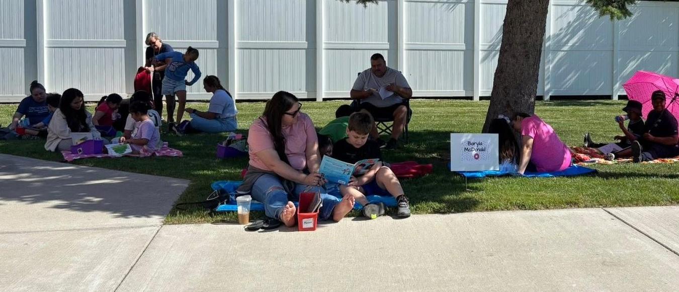 Families sit on blankets in the grass reading books outside under a large pine tree.