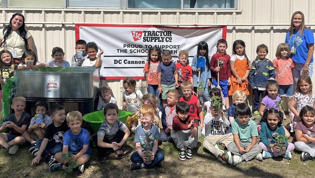 A diverse group of children and adults smiling in front of a school garden banner.